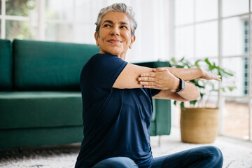 Happy and healthy senior woman doing a cross arm stretch in a peaceful yoga session at home - obrazy, fototapety, plakaty
