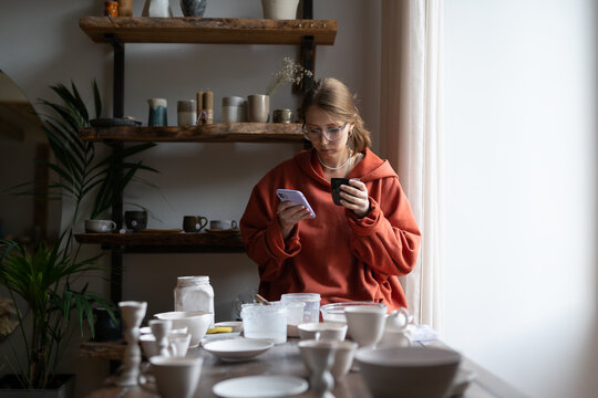 Craftswoman with phone sits in pottery workshop photographs handmade clay products. Girl artisan with smartphone takes photo of tableware for sale through social networks. Take a break from work