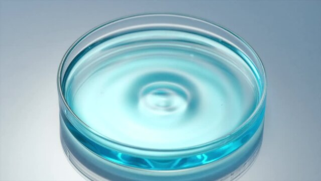 Close up scene of water drop dropping on surface of blue liquid on petri dish, create small waves and bubbles in slow motion. Concept of medicine or cosmetic research and organic skin or body care pro