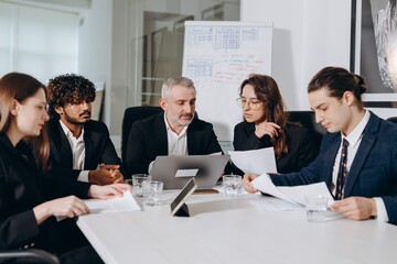 A team of businessmen led by a middle-aged man review documents during a meeting. Businessmen look through reports for the last financial quarter.