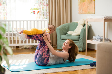 Young mother working out at home on a mat with baby. Interior of a cozy sunny nursery room. Woman practicing yoga together with child.