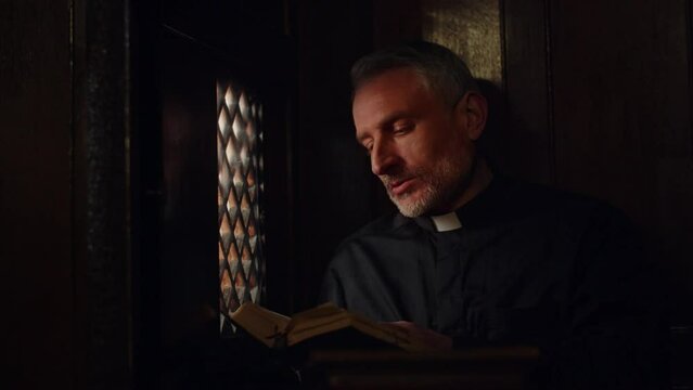 The priest listens to confession while sitting in the confessional. A pastor in a black robe is quoting a passage from the Bible to a man.