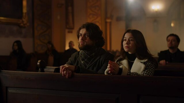 A brunette man and his girlfriend are sitting in a church during the Sunday service. Husband and wife close their eyes and fold their hands in prayer.