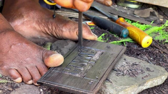 Local Tanzanian man holding a stone plate with toe fingers and carving the ANNA word using a metal chisels and hammer. Traveling and craftsman's everyday work 4K concept footage.
