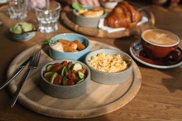 Breakfast table with scrambled eggs, sausages, avocado, croissant, cucumber, tomato, coffee and water