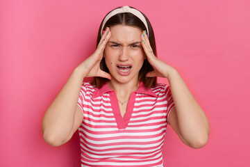 Obraz na płótnie Canvas Indoor shot of irritated angry aggressive young girl with hair band wearing striped casual t shirt standing isolated pink background, screaming with angry, having headache