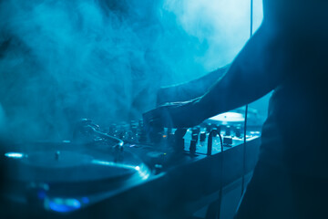 Club DJ mixing vinyl records in blue stage lights and thick smoke. Disc jockey playing music in...