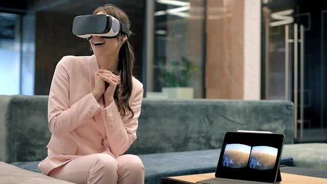 A blonde caucasian excited woman exploring virtual reality using VR glasses in an office. Tablet on the table showing image visible in the headset