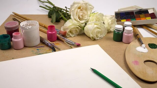 A bouquet of white roses and painting supplies are located on the artist's desk. A wooden palette, a variety of brushes and paints, paper and pencils are on the table.