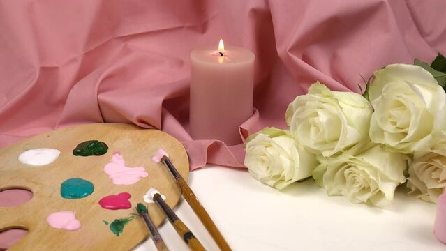 Composition for drawing from life of a pink burning candle, paint cans, wooden palette for paints, paint brushes and white fresh roses against the pink textile background in an art school 