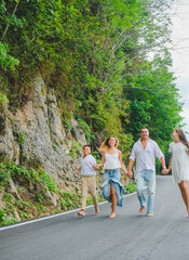 Cheerful, happy family, father, mother, son and daughter enjoying walk play outdoor near sea. Funny family in white clothes barefoot running on walkway. Green rocks at background.