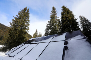 Solar panels covered with snow nearby of a mountain cottage in a beautiful winter day. Photovoltaic solar energy eco friendly technology system industry.