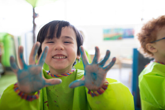 Playful preschool students in smocks showing finger paint on hands at poster in classroom