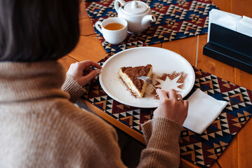 Unknown woman eating tiramisu cake on white plate standing at wooden table