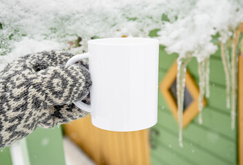 Traditional pattern mitten holding white blank coffee mug mock up template. Cozy seasonal products advertisement background. Single cup on snowy Scandinavian winter nature background. Room for text.