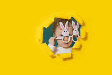 Little surprised baby boy in bunny shape glasses looking, peeping through the yellow paper hole....