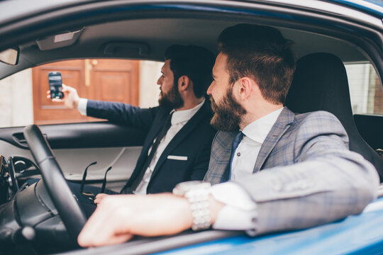 Two elegant contemporary bearded businessmen taking selfie seated in car