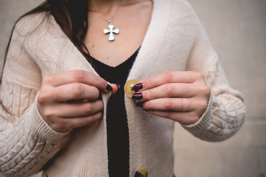 Closeup of religious girl with jewelry cross, beautiful hands, dark nails and beige sweater (with fabric details) in sunny city