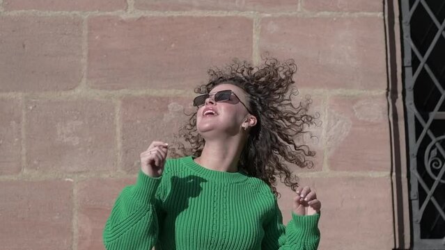 Dancing fashionable young woman with curly hair in sweater and dark sunglasses has fun in the sunny city