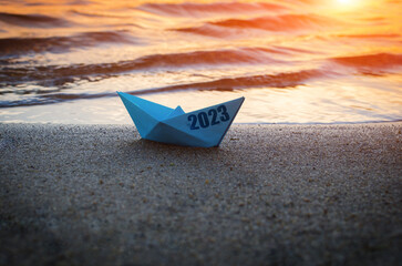 Blue paper boat with inscription 2023, on sandy shore beach against sunset.