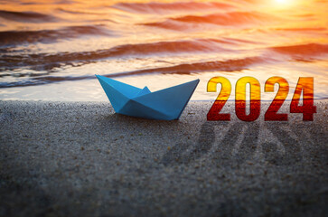 Blue paper boat and year 2024 on sandy beach.Against background sunset.