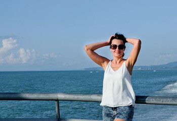 Positive woman walking and enjoys sea vacation and sunshine. Cheerful, smiling, lucky woman in white blouse, shorts and sunglasses, outdoors on the pier in Batumi, Georgia.