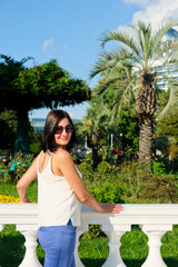Nice woman walk at city enjoys vacation. Flowers garden with palm at background. Cheerful, smiling, lucky woman in white blouse, blue trousers and sunglasses outdoors. Batumi, Georgia.