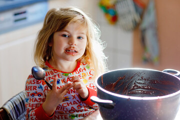 Cute toddler girl eating chocolate dough rests with spoon and fingers from pot. Happy child licking sweet dough for muffins or cake, helping in home kitchen, indoors. - obrazy, fototapety, plakaty