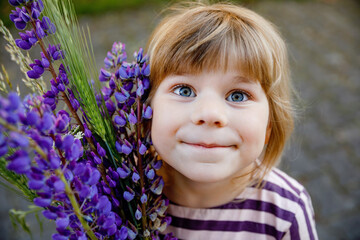 Cute adorable little toddler girl with huge bouquet of blossoming violet lupnies flowers. Portrait of smiling preschool child in domestic garden on warm spring or summer day. Summertime.