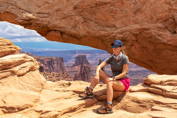 Woman sitting in front of Mesa Arch near Moab, Canyonlands National Park, San Juan County, Southern Utah, USA. Looking at natural pothole arch rock formation on eastern edge of Island in the Sky Mesa