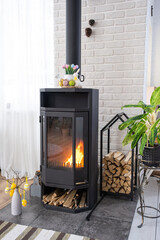 Easter decor near fireplace stove with fire and firewood. Cozy home hearth in interior with potted plans, colorful easter eggs, easter bunny and bouquet. Spring in a country house