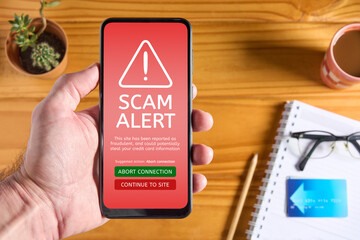 Close up of male hand holding mobile phone with red screen and warning icon with scam alert text and message saying that the site could steal credit card information. Accessories in the background - obrazy, fototapety, plakaty