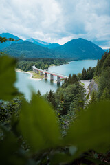 Kurvige Brücke und Straße über einen See mit Bergen und Wolken und schöner Natur und...