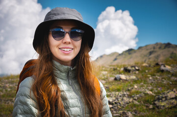 Portrait of a traveler in the mountains. The concept of adventure, travel and hiking. Happy woman in a cap enjoys the sunshine while hiking in the mountains. Tourist with glasses