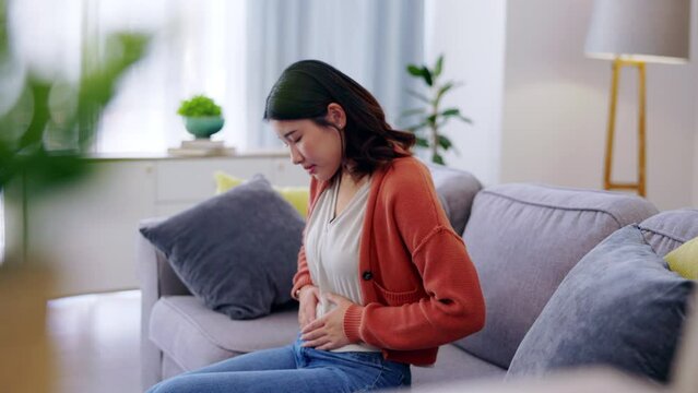 Pain, sick and a woman feeling stomach for ibs, problem and insecurity. Sad, depression and Asian girl touching her belly for body dysmorphia, illness and uncomfortable gas on the lounge sofa
