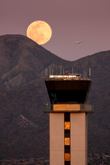 This large full moor rises over the McDowell Mountaind range and the airport tower at Scottsdale, Arizona Airport. A business jet on approach to the airport is visible close to the full moon. 