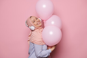 Fototapeta na wymiar Attractive glamour Middle-Eastern Muslim woman in a pink hijab and wireless headphones, having fun on her birthday party, listens to music, smiles, poses with pastel color balloons on pink background