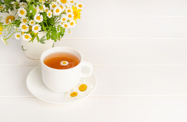 Chamomile tea in a white cup on a white wooden background with a bouquet of daisies with copy space.