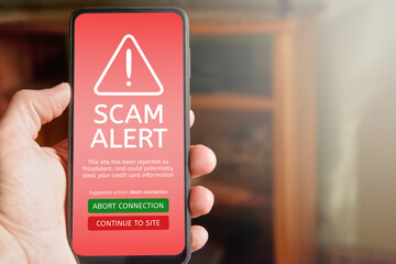 Close up of male hand holding mobile phone with red screen and warning icon with scam alert text...