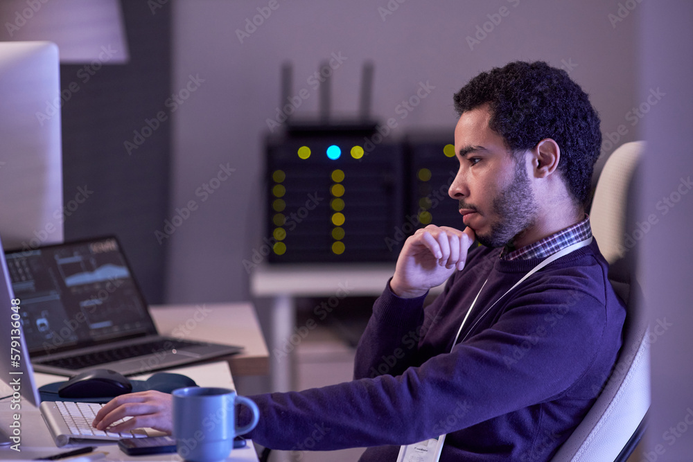 Poster side view portrait of software developer working at night writing code at desk in office lit by neon