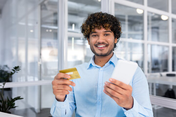 A young hispanic man is sitting in the office, holding a phone and using a credit card. Smiling...