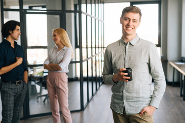 Portrait of positive young male office worker in casual clothes holding takeaway coffee cup in hand standing in office, smiling looking at camera. Startup business team friendly chatting on background