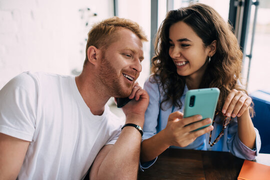 Cheerful Caucasian male and female bloggers reading funny content publication during friendly meeting in cafe interior, happy couple in love using cellphone gadgets for social networking and calling