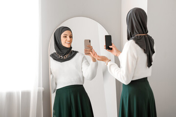 Beautiful Muslim woman with mobile phone taking selfie in front of mirror at home