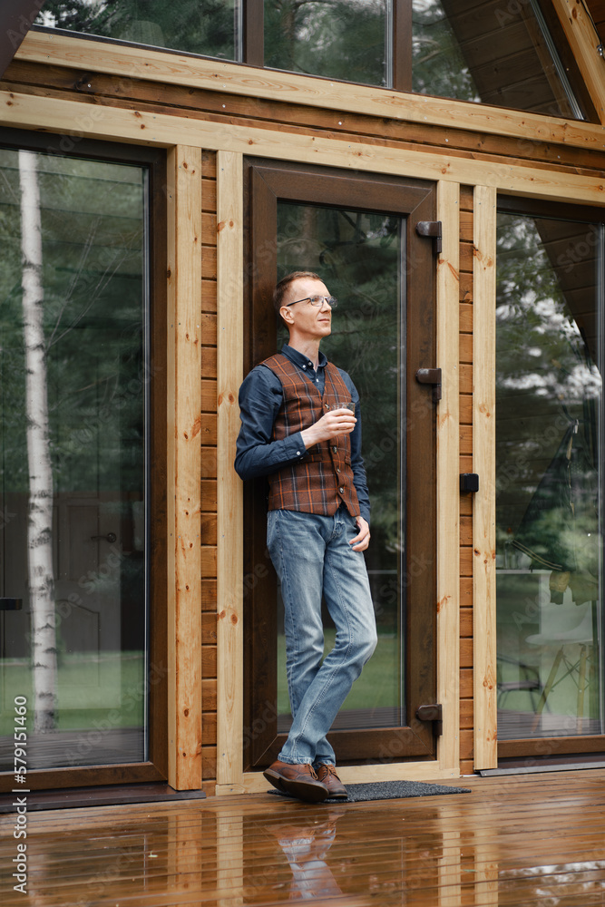 Wall mural pensive middle age man stands on the terrace of a wooden bungalow and drinks whisky