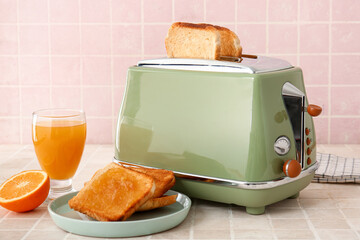 Bread slices in modern toaster, cup of juice, orange and toasts with jam on tile table