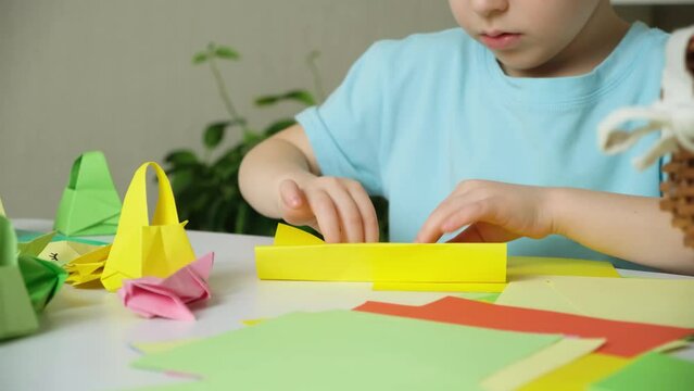 Preschool boy with rabbit ears nag head makes Easter crafts, origami figurines of rabbits and baskets of paper