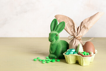 Bunny, holder with Easter gift bag, chocolate egg and candies on green table against beige wall