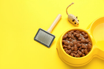 Bowl of wet pet food, toy and grooming brush on yellow background
