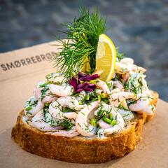 Close-up of a traditional Danish delicacy sandwich, adorned with prawns, dill, chives, lemon slices, and edible flowers, with the lettering smørrebrød, engl. butter and bread, in the background.